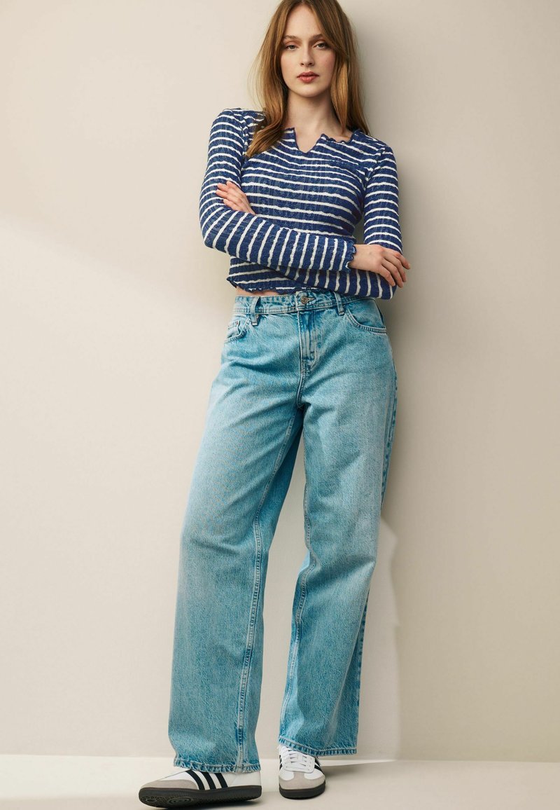 Helle blaue, weit geschnittene Jeans aus Denim, kombiniert mit einem langärmeligen Oberteil in Marineblau und Weiß gestreift. Graue und weiße Turnschuhe vervollständigen das Outfit.