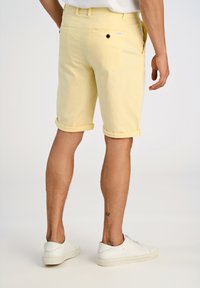 Shorts en coton jaunes avec revers, poche arrière simple avec fermeture à bouton, portés avec des baskets blanches. Design simple.