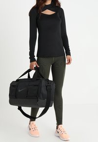 Top sportivo nero a maniche lunghe con dettagli a taglio, leggings testurizzati verde oliva e borsa da palestra nera Nike con più tasche. Sneakers rosa.