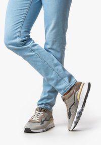 Sneakers con una combinazione di suede grigio e marrone e mesh, dotate di lacci bianchi e una suola strutturata in bianco e nero. Indossati con jeans azzurri chiari.