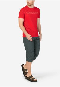 Rotes T-Shirt mit kurzen Ärmeln und "TIMEZONE & CO" Grafik, kombiniert mit knielangen dunkelgrauen Shorts mit strukturiertem Muster und bequemen braunen Sandalen.