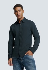 Donkergroene longsleeve shirt met een knoopsluiting aan de voorkant, kraag en aangesloten mouwen, gecombineerd met blauwe spijkerbroek. Gladde stoftextuur en casual ontwerp.