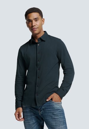 Chemise à manches longues vert foncé avec un devant à boutons, un col et des manches ajustées, associée à un jean en denim bleu. Texture de tissu lisse et design décontracté.