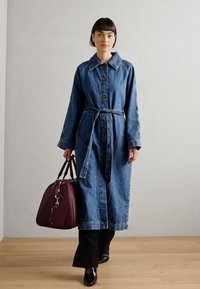 Denim trench coat with a belt, blue in color, featuring a collared neckline and button front. Paired with a burgundy handbag and black flared pants.