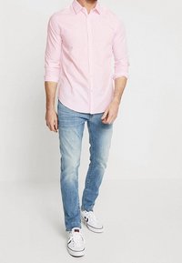 Camicia rosa chiaro a maniche lunghe con bottoni abbinata a jeans blu chiaro e sneakers bianche. Il tessuto appare liscio, con una vestibilità rilassata.