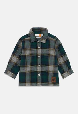 Chemise en flanelle à carreaux verts et gris avec des manches longues, un col pointu et des boutons blancs ; elle présente un logo en cuir marron en bas.
