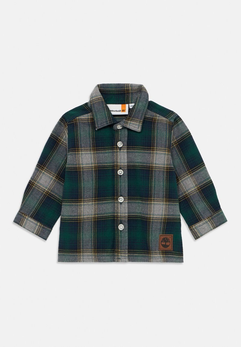 Chemise en flanelle à carreaux verts et gris avec des manches longues, un col pointu et des boutons blancs ; elle présente un logo en cuir marron en bas.