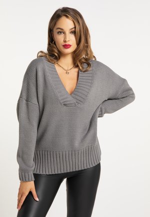 Femme aux cheveux bouclés porte un pull en maille gris à col en V et un pantalon en cuir noir, posant avec une main sur la hanche sur fond blanc.