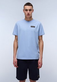 Lyseblå bomuldstrøje med rund hals, korte �ærmer og et rektangulært logo-patch på brystet. Kombineret med mørke shorts.