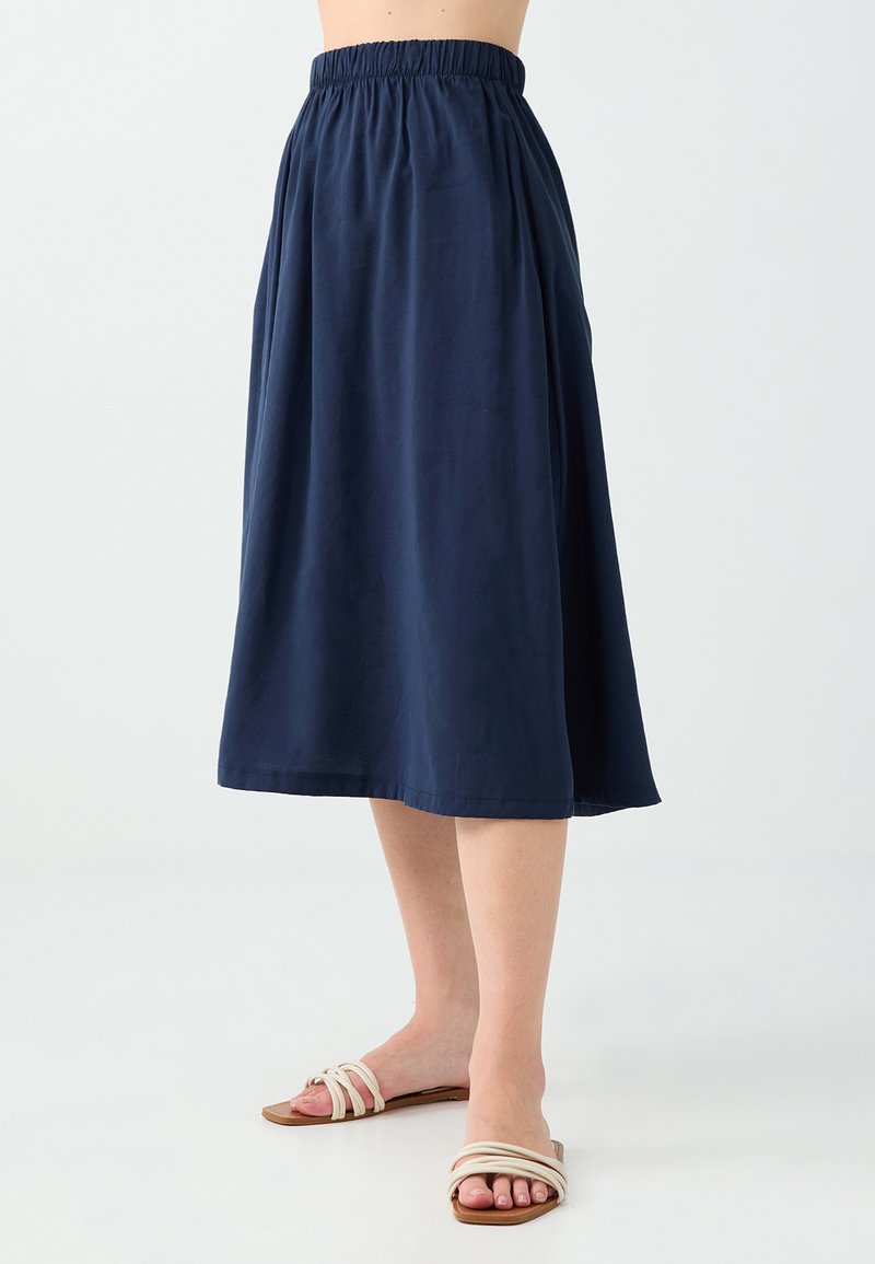 Femme portant une jupe midi bleu marine à taille élastique et des sandales blanches plates à trois brides, debout sur un sol blanc.