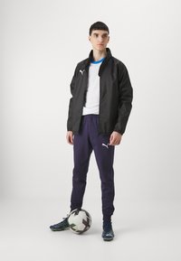 Black windbreaker with logo, white T-shirt, purple joggers, and blue football boots. Holding a black-and-white soccer ball.