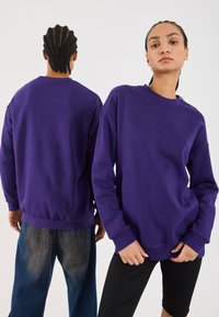 Twee mensen dragen paarse sweaters, de een kijkt naar voren met de handen naast de heupen, de ander kijkt achterover en toont een gevlochten haarstijl.