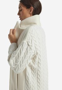 Cappotto bianco oversize con maniche in maglia a trecce testurizzate, colletto alto e tessuto morbido e liscio. Presenta una vestibilità rilassata e una palette di colori neutra.