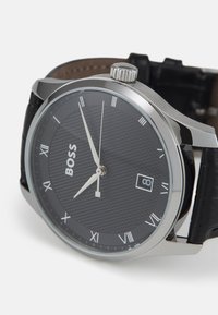 Reloj de acero inoxidable tono plateado con un dial texturado negro, números romanos y una ventana de fecha. La correa de cuero negro completa el diseño.