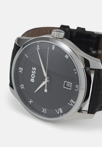 Montre en acier inoxydable argenté avec un cadran noir texturé, des chiffres romains et un affichage de la date. Un bracelet en cuir noir complète le design.