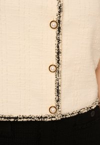 Béžový tweedový top s krátkým rukávem, zdobený černým lemováním a akcenty zlatých knoflíků s perlovými detaily na přední straně. Texturovaná tkanina.