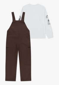 Pruunid puuvillased üleüldised riided reguleeritavate rihmadega ja kahe esikotiga, kombineeritud valge pika varrukaga särgiga, millel on graafilised trüki detailid varrukatel.