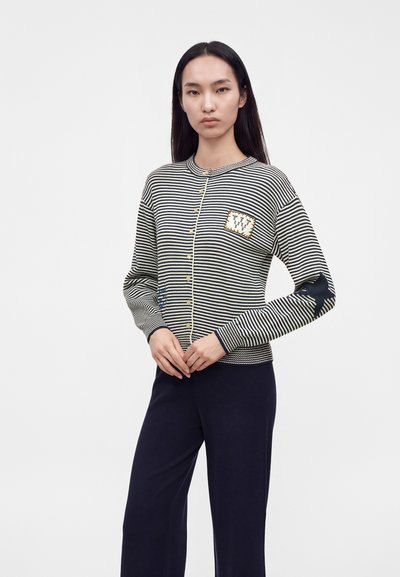 Femme aux longs cheveux foncés porte un pull boutonné rayé noir et blanc avec un écusson et un pantalon noir, debout devant un fond uni.