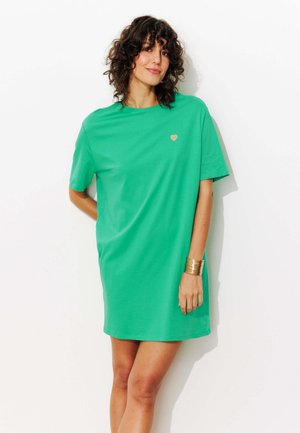 Femme aux cheveux bouclés portant une robe verte ample à manches courtes avec un petit cœur doré et un bracelet manchette doré, debout contre un mur blanc.