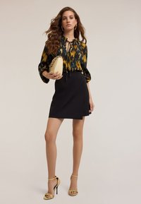 Falda negra envolvente combinada con una blusa de estampado multicolor con cuello de lazo. Acentuada con sandalias de tacón amarillo y un bolso pequeño.