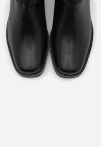 Bottines noires en cuir avec une forme de pointe, texture lisse et coutures visibles le long des bords. Design minimaliste, sans embellissements.