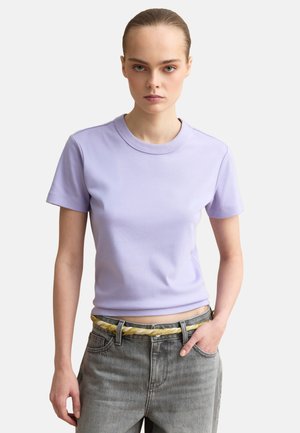 Giovane donna con maglietta a maniche corte viola chiaro, jeans grigi e cintura gialla intrecciata, in piedi con una mano in tasca su sfondo bianco.