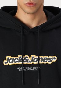 Fekete kapucnis pulóver zsinórral, elülső részén feltűnő fehér és sárga "Jack & Jones" logóval. Puha szövet tapintás.