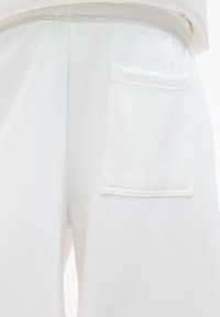 Pantalon de survêtement blanc en tissu doux, doté d'une taille élastique et d'une poche plaquée à l'arrière avec des coutures visibles.