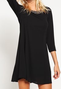 Robe noire en tissu doux, avec manches trois-quarts, coupe décontractée, et détail de col orné de perles. Texture lisse et silhouette fluide.