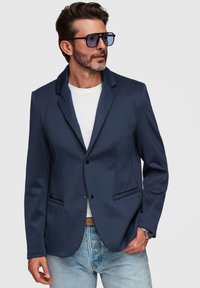 Homme portant un blazer bleu foncé, une chemise blanche, un jean bleu clair, des lunettes de soleil et une montre, posant devant un fond gris clair.