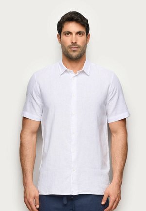 Mężczyzna o krótkich ciemnych włosach, ubrany w białą koszulę z krótkim rękawem i ciemnoniebieskie spodnie, stojący na jasnym jednolitym tle.