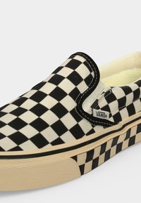 Μαύρο και άσπρο καρό παπούτσι Vans τύπου slip-on με λαστιχένια σόλα και λογότυπο στην πλαϊνή ετικέτα σε ανοιχτό φόντο.