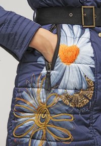 Abrigo acolchado azul marino con bordado floral en azul, dorado y naranja, que cuenta con un bolsillo tipo cremallera lateral y un cinturón negro en la cintura.