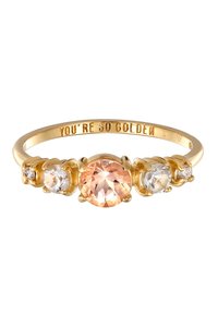 Guldfärgad ring med en central rund persikoädelsten flankeras av två klara stenar, med graverad text på insidan av ringbandet.