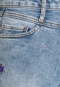 Jeans de mezclilla azul claro con un acabado desgastado y bordados florales multicolores en el bolsillo trasero. Costuras amarillas que realzan el diseño.