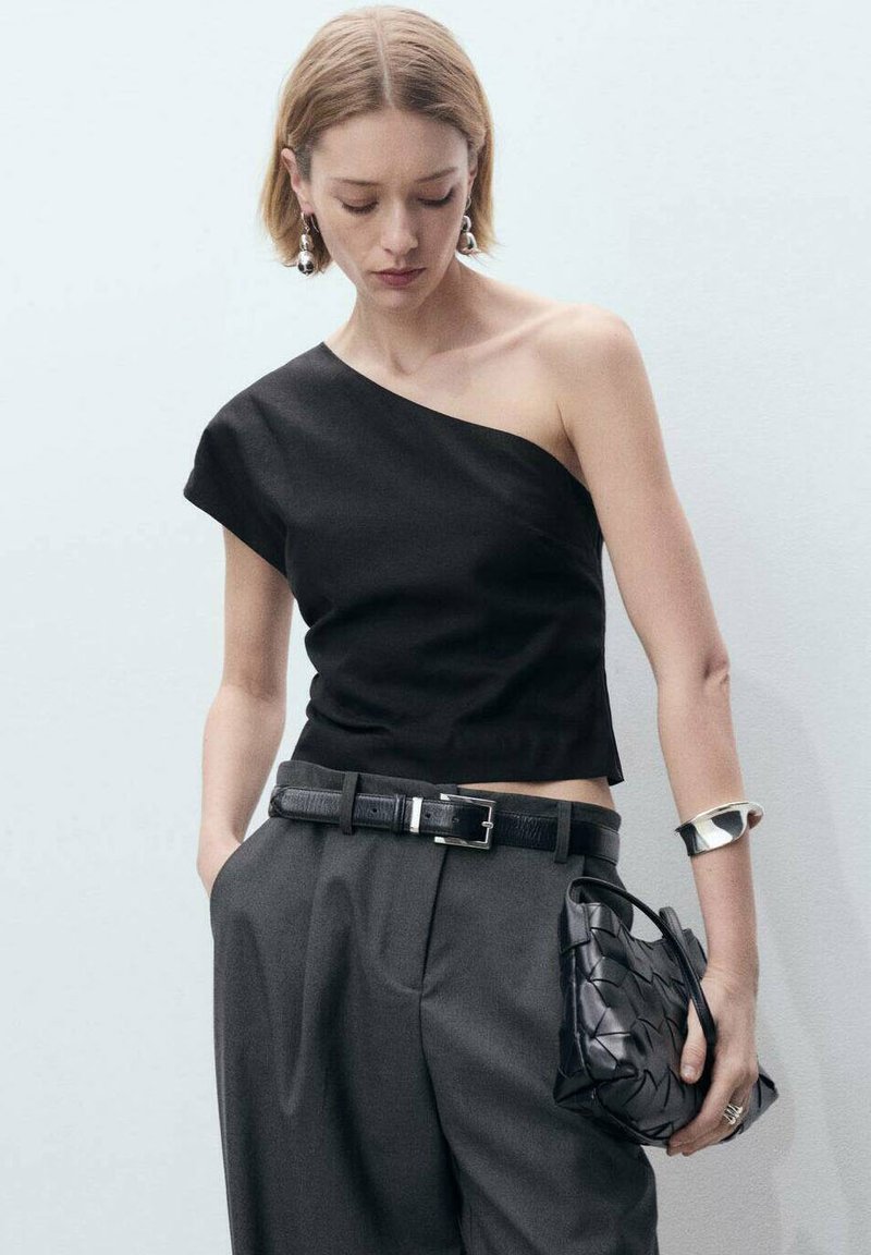 Top nero a spalla unica con un design cropped, abbinato a pantaloni grigi a vita alta, una borsa intrecciata nera e accenti di gioielli argentati.