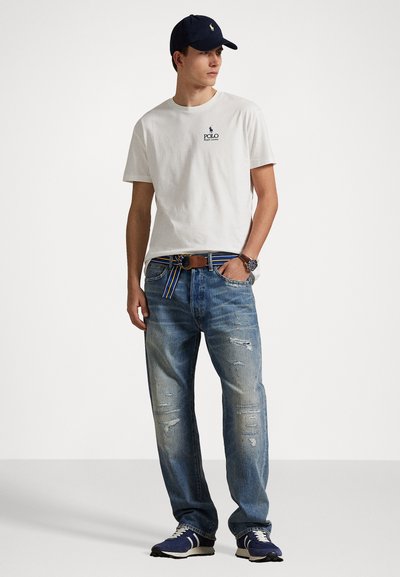 Tricou alb din bumbac cu un logo bleumarin, blugi decolorati, și o șapcă bleumarin. Ținuta este completată cu o curea multicolorată cu dungi și adidași.