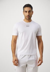 Camiseta deportiva blanca hecha de una tela suave y ligera. Presenta el logotipo de EA7 en el pecho y un sutil branding a lo largo de los lados. Mangas cortas.
