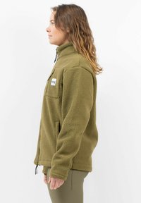 Olivgrüne Fleecejacke mit hohem Kragen, Reißverschluss-Tasche und kontrastierendes weißes Etikett. Hat eine lockere Passform und texturierten Stoff.