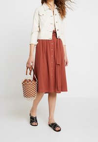 Woman in a cream cropped jacket and rust button-front midi skirt holds a netted tote bag, wearing black studded slide sandals.