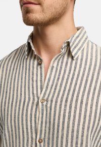 Camicia a righe con bottoni, caratterizzata da sottili strisce blu navy e crema, realizzata in un tessuto morbido con colletto e bottoni di legno. Finitura testurizzata visibile.