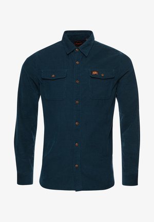 Homme marchant à l'intérieur portant une chemise boutonnée vert sarcelle foncé, un pantalon vert olive, des bottes noires et des bracelets à son poignet gauche.