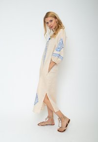 Hellbeiges Leinenkleid mit blauer Blumenstickerei an den langen Ärmeln und einem offenen Vorderteil, mit Seitenschlitzen und kombiniert mit beige farbigen Sandalen mit Riemchen.