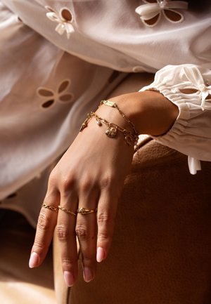 Gouden sieraden op een hand, met gelaagde armbanden met bedels, een gestructureerde ring en gladde nagels, tegen een neutrale achtergrond.