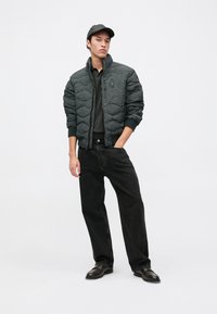Jacket bombers înfășurat de culoare gri închis, cu guler înalt, manșete ribbed, fermoar frontal și un buzunar pe piept, purtat cu blugi negri largi și pantofi negri.