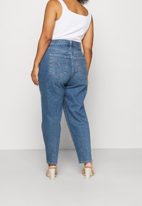 Modré džínové kalhoty s vysokým pasem a zúženými nohavicemi, s kapsami na zadní straně a výraznou logovou nášivkou, doplněné béžovými sandály na podpatku.