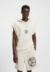 Crème colored mouwloze hoodie met een voorzak en een ronde graphic op de borst, gecombineerd met bijpassende shorts met een groot bedrukt logo.