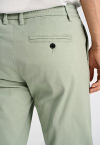 Pantaloni verde chiaro con una texture liscia, dotati di una tasca posteriore con un bottone nero e passanti per la cintura. Design semplice e funzionale.