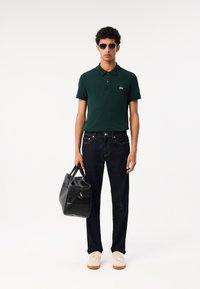 Camisa polo verde oscuro, jeans oscuros, zapatillas beige, sosteniendo un bolso negro texturizado, complementado con gafas de sol. Diseño simple y funcional.