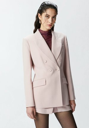 Blazer doppiopetto rosa pallido con rever a rotondo, due tasche frontali e pantaloni abbinati. Indossato sopra un top bordeaux a pois.